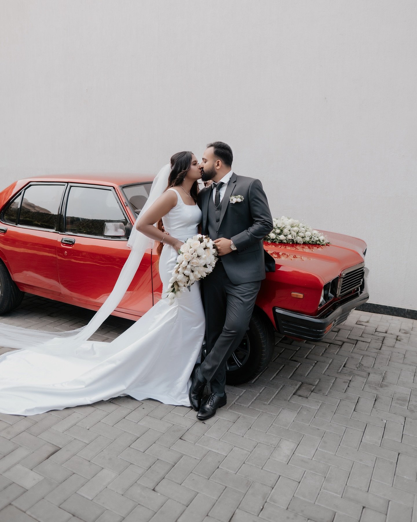 A newlywed couple kissing beside a flower-decorated red vintage car